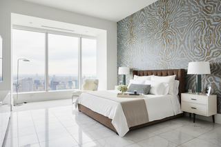 A modern bedroom interior featuring a large bed with white bedding and brown upholstered base and headboard, set against a patterned accent wall. The room has a large floor-to-ceiling window with a city view, white nightstands with lamps, a light chair, and polished tile flooring.